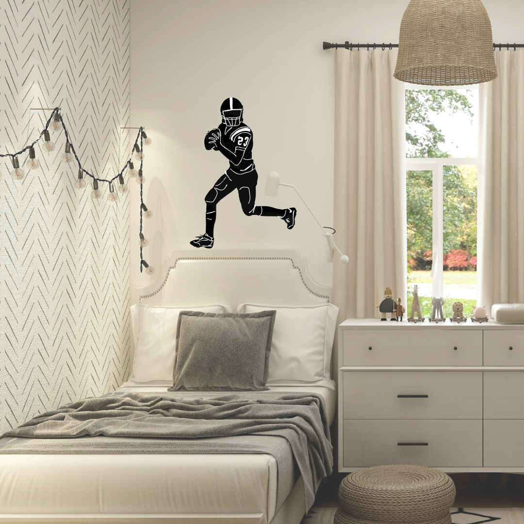 A custom black vinyl wall decal of a football player in a running pose is displayed on a bedroom wall above a bed. The room features a white bed with gray bedding, a white nightstand, a woven pendant light, and a window with beige curtains.