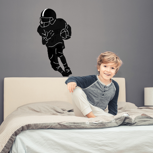 A young boy sitting on a bed in a bedroom with a custom vinyl decal of a football player on the gray wall above him.