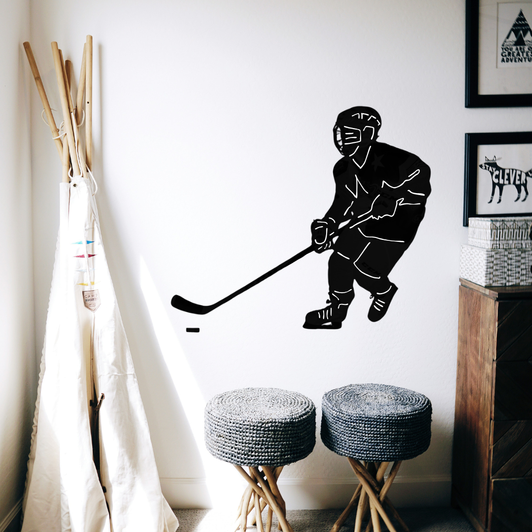 A custom black vinyl wall decal of a hockey player displayed on a white wall in a styled room setting with two woven stools, a wooden coat rack, and framed wall art.