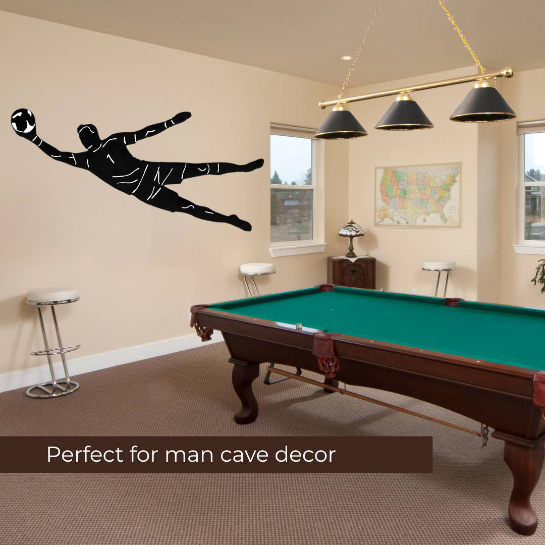 Custom-made wall decal of a soccer goalkeeper diving to catch a soccer ball, shown on a wall in a man cave setting with a pool table, bar stools, and pendant lights. A text banner at the bottom reads 'Perfect for man cave decor'.