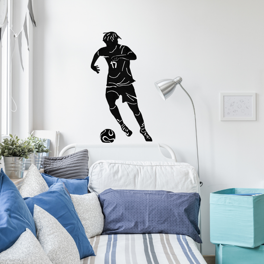 Custom silhouette vinyl wall decal of a soccer player displayed on a white bedroom wall above a bed with blue and white pillows.