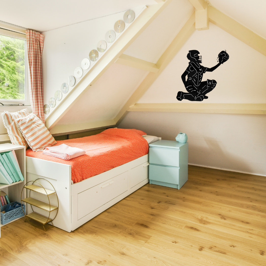 A black silhouette wall decal of a softball catcher displayed on the slanted wall of an attic bedroom above a light blue nightstand and a bed with an orange blanket.